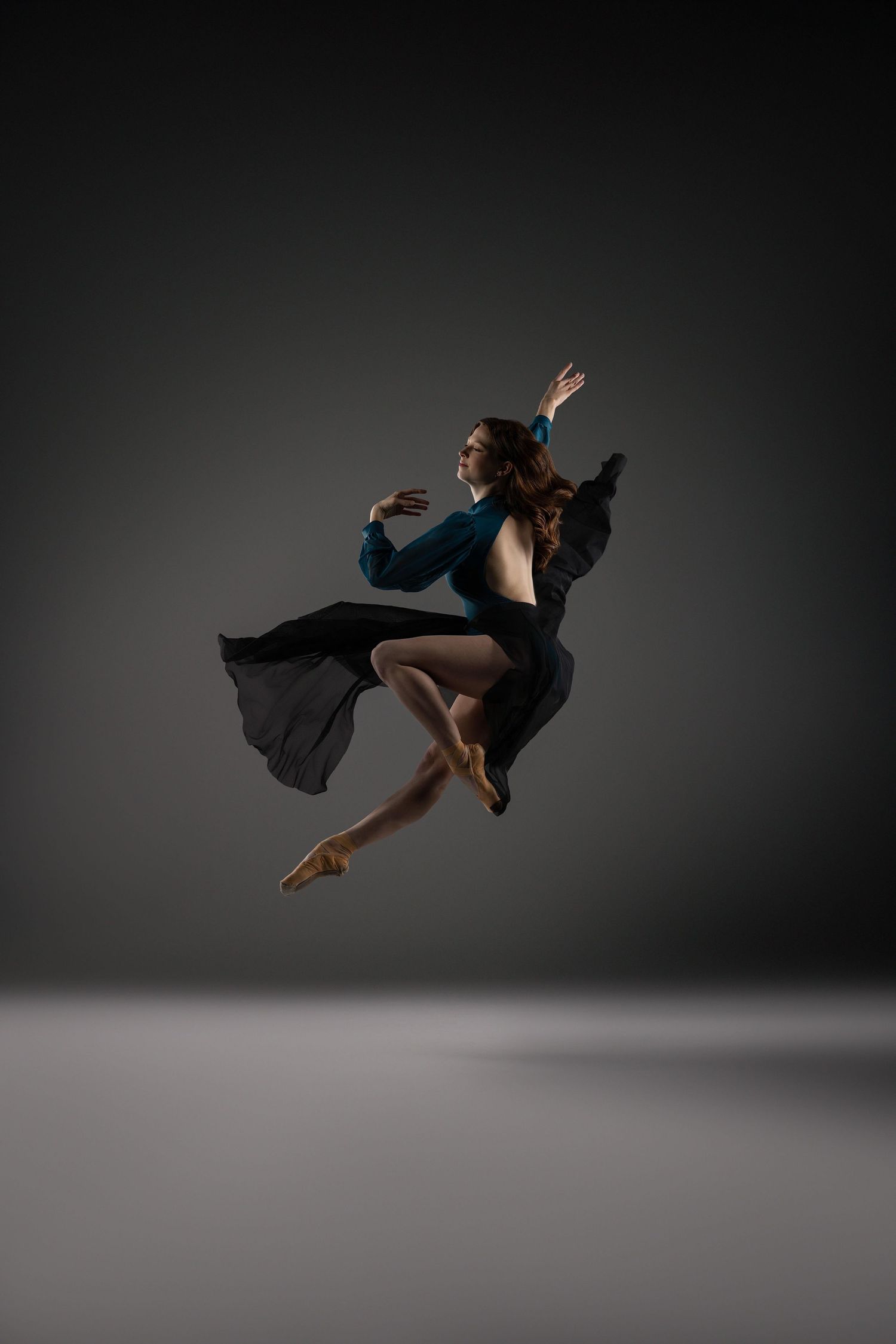 Dancer in flowing black costume leaps gracefully through the air against dark background in dramatic studio lighting.