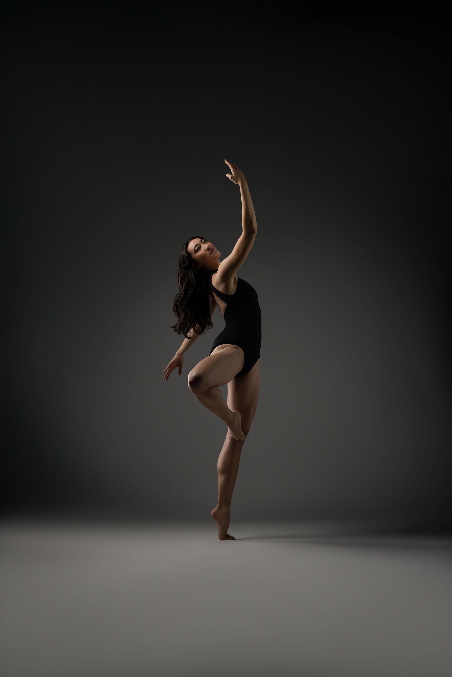 Dancer in black leotard performs elegant ballet pose against dark background in dramatic studio lighting.