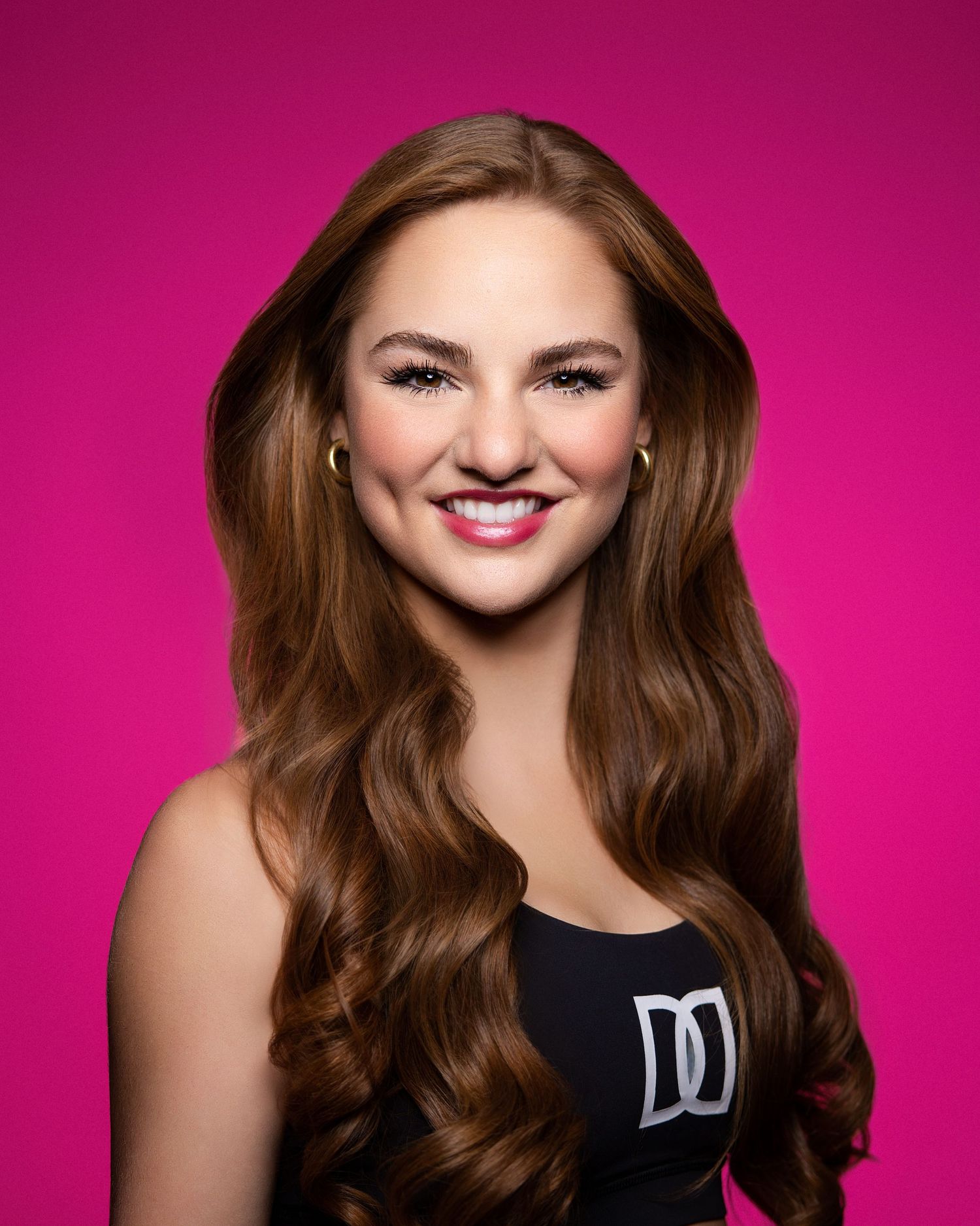 Professional headshot portrait against a bright pink background showing a warm smile and long wavy brown hair.