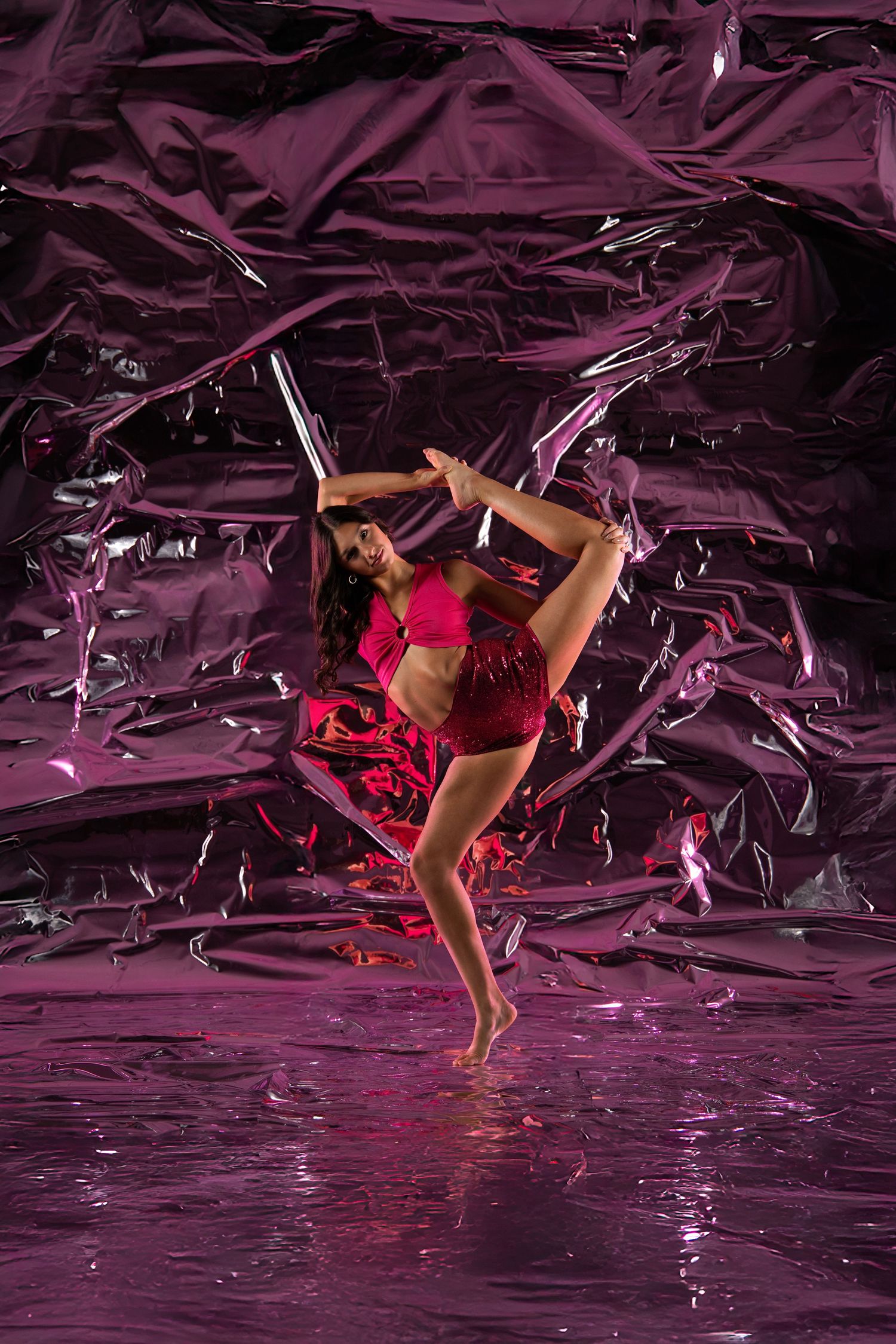 A dancer in a pink sequin outfit performs a high kick against a shiny crumpled metallic pink background.