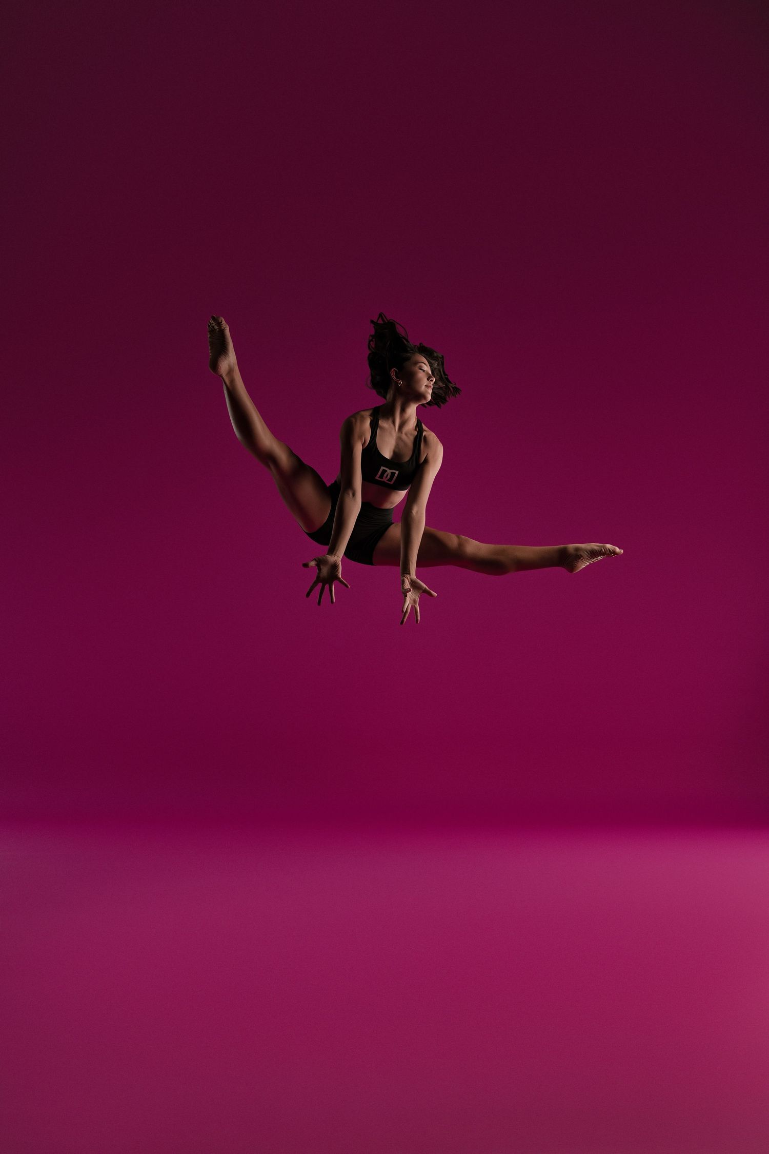 Athletic dancer in black leotard performs dramatic split leap against dark magenta background. From Dance Dynamics, Texas.