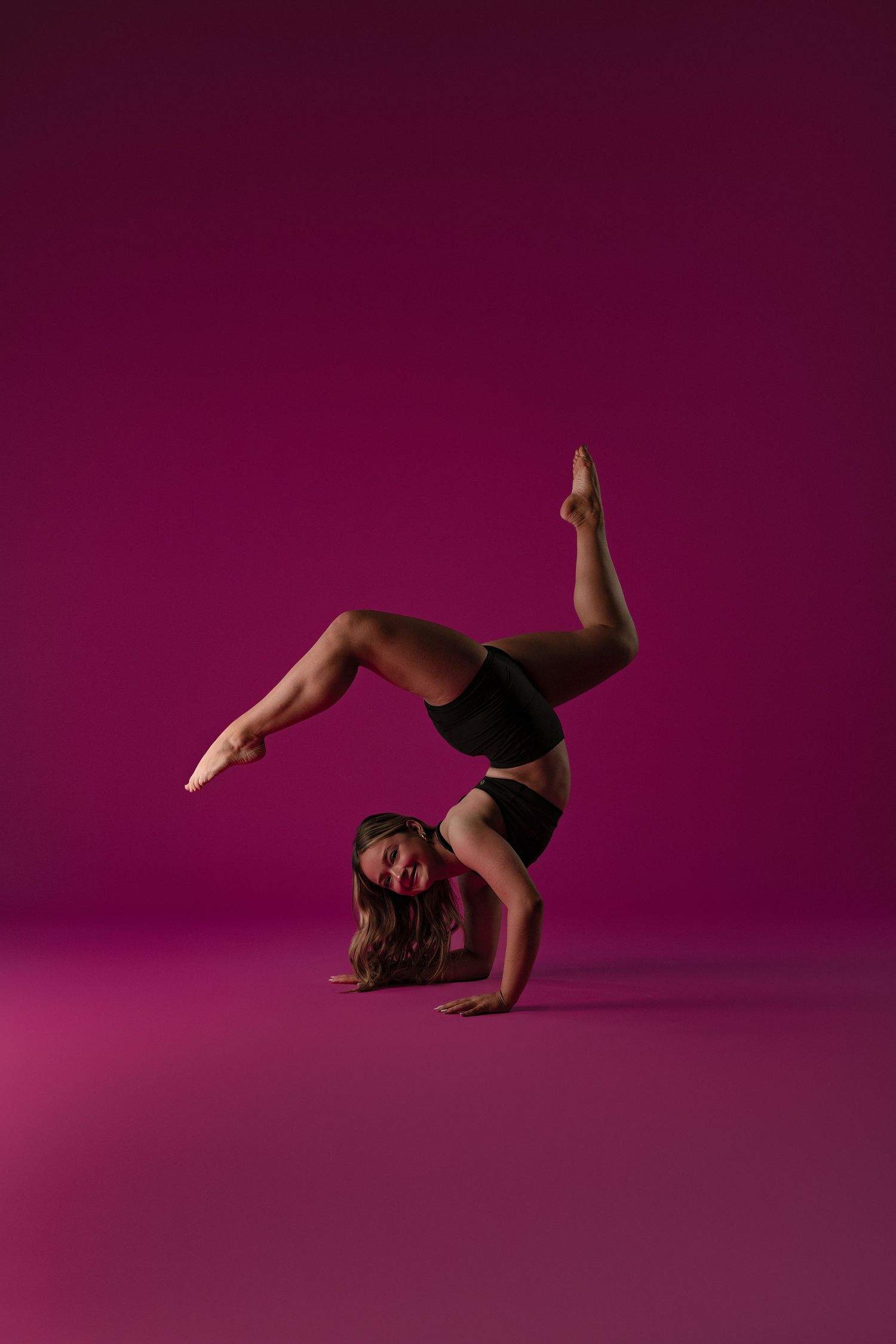 Skilled gymnast performs a graceful forearm stand pose against a deep purple background, wearing black athletic wear.