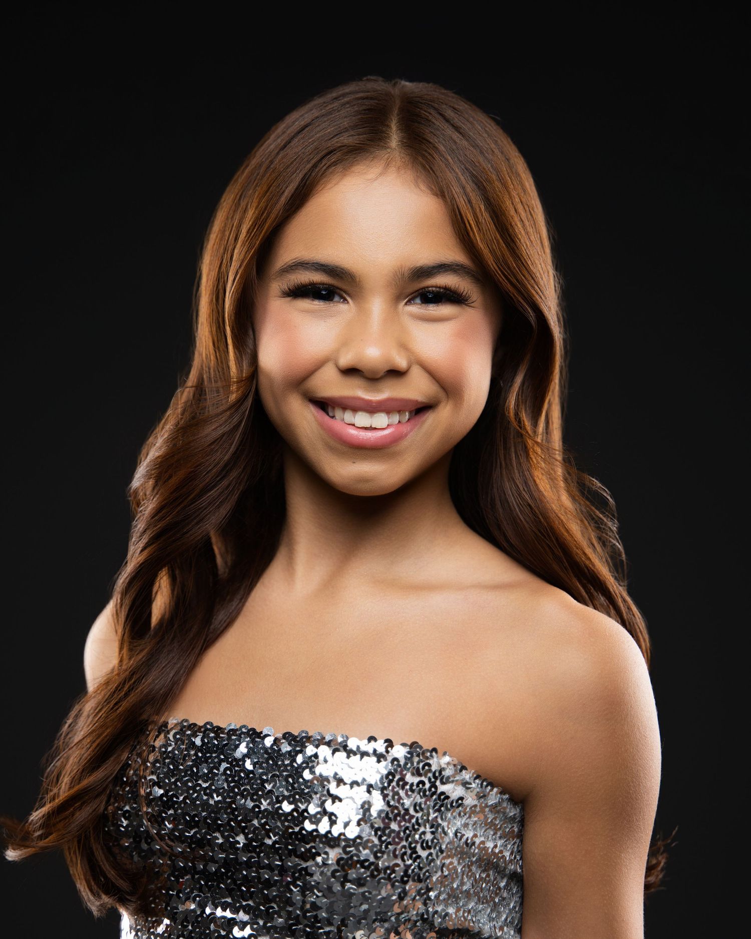 Young girl smiling in a silver sequin strapless top with long brown hair against dark background.