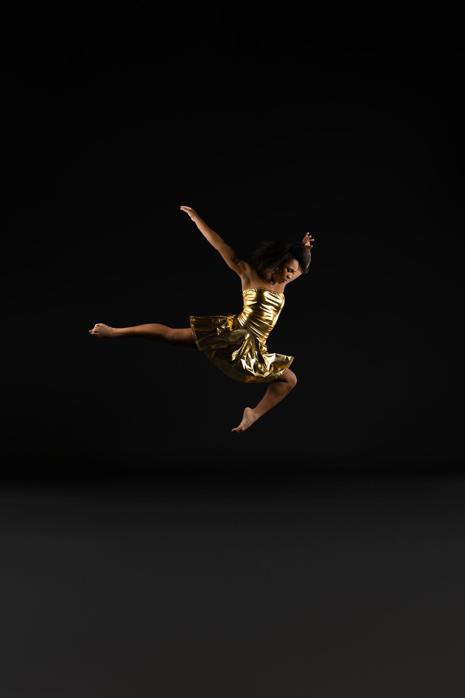 A dancer in a gold dress leaps gracefully against a dark background, arms extended in an elegant pose.