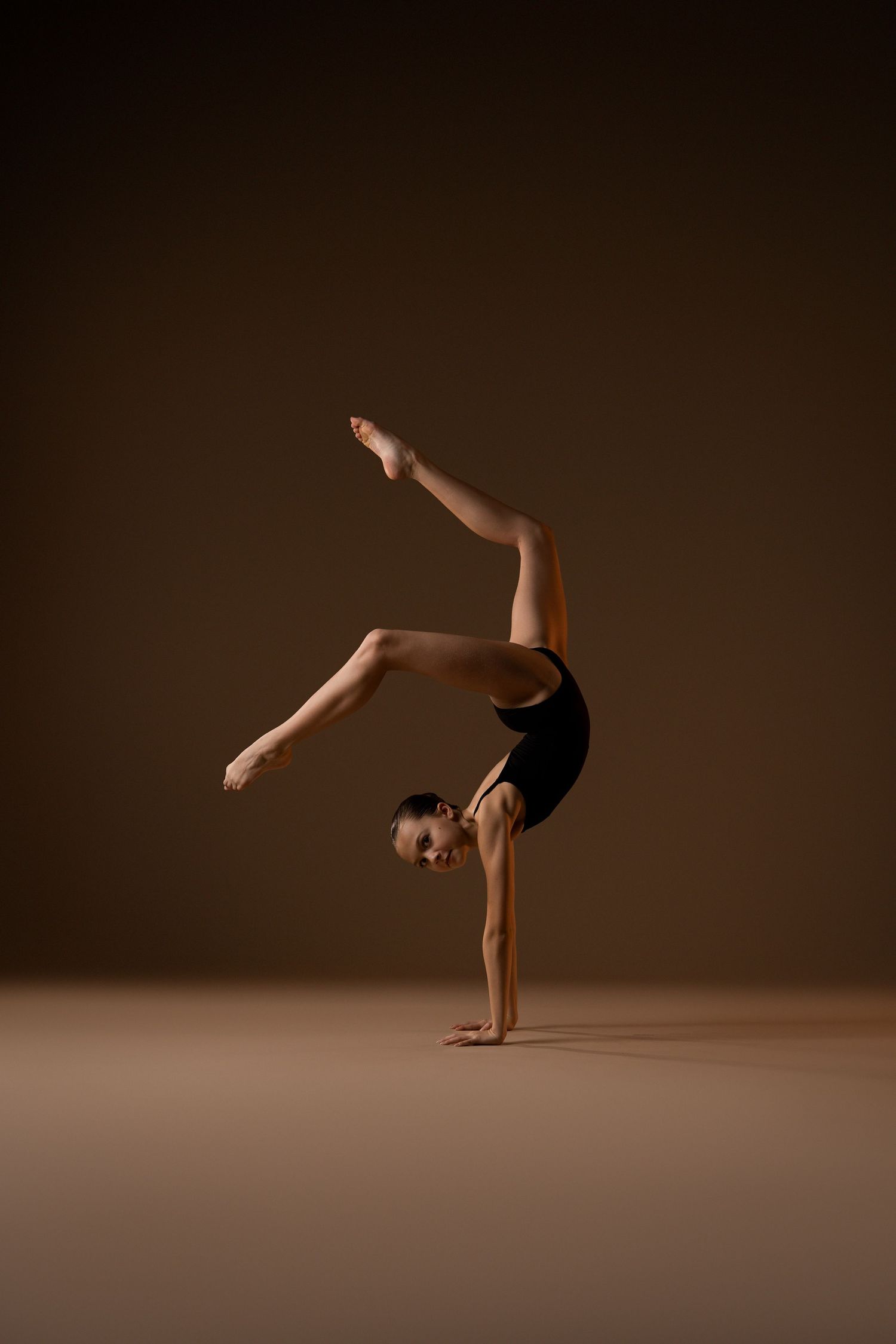 A gymnast in a black leotard performs a one-handed handstand with legs split on a brown studio background.