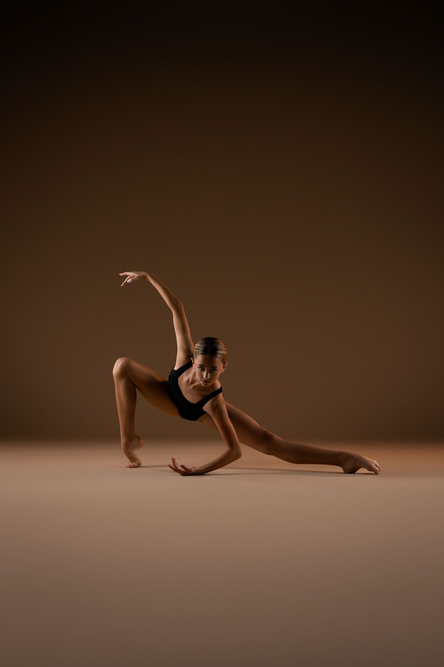A flexible dancer in a black leotard performs a side split stretch pose against a warm brown studio background.