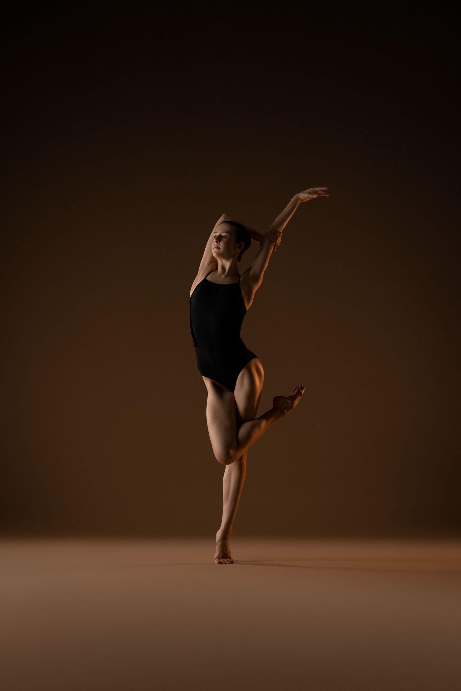 A graceful ballerina in a black leotard performs an elegant pose on one leg against a warm brown studio background. Woodlands Ballet Academy