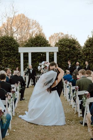 Outdoor wedding celebration with white pillared arch and gathered guests on lawn.