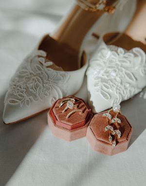 White lace wedding shoes with rose gold ring boxes on white fabric.