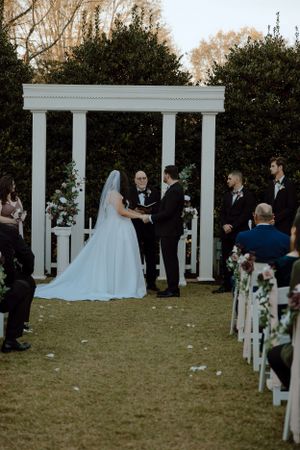 An outdoor wedding ceremony under white columns with guests seated on the lawn.