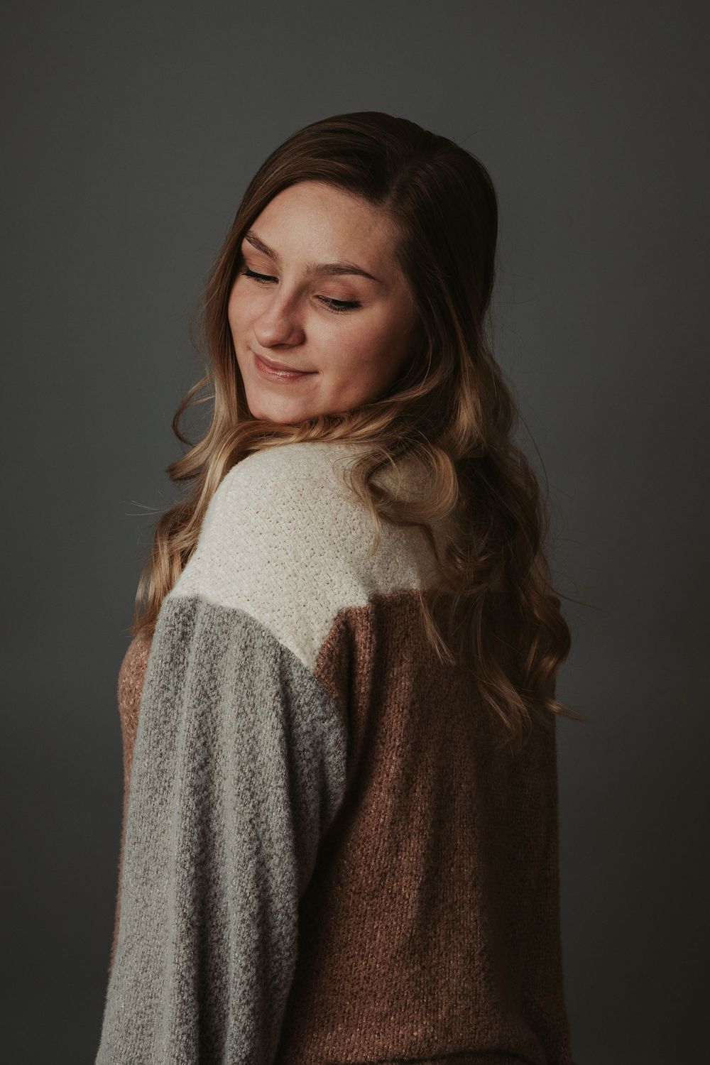 Portrait of someone in a cozy color block sweater looking down with a gentle smile against a dark background.
