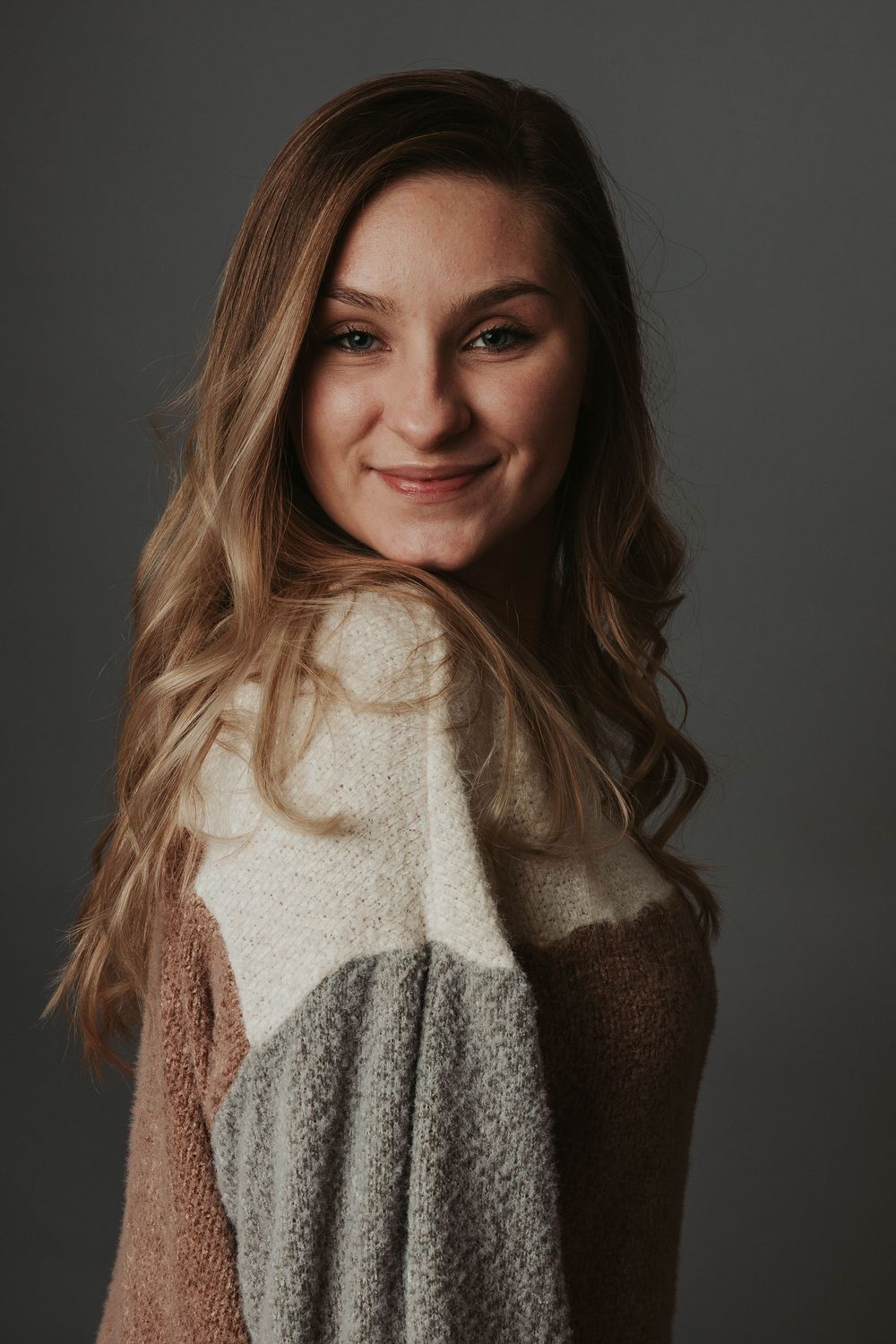 Studio portrait showing a warm smile while wearing a soft tricolor sweater against grey backdrop.