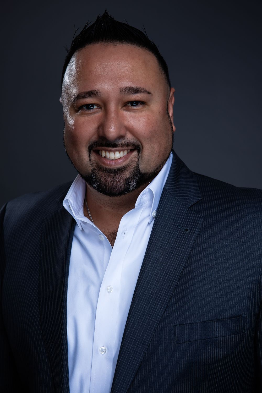 Professional headshot portrait of a smiling man in a dark suit jacket and white dress shirt against a dark background.