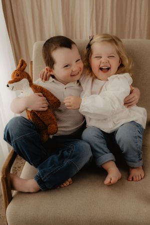Two kids sharing joyful laughter while holding stuffed fox toy on beige chair.