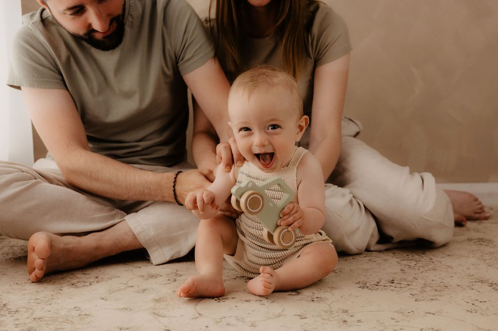A happy baby wearing a neutral onesie sits on the floor with two people in casual attire behind them.