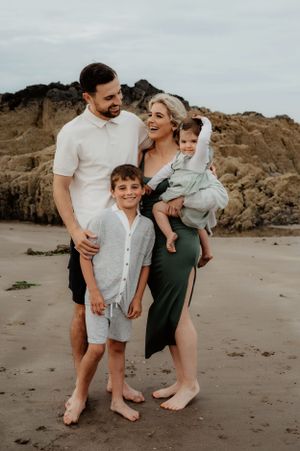 A family poses together barefoot on a beach with rocky cliffs in the background.