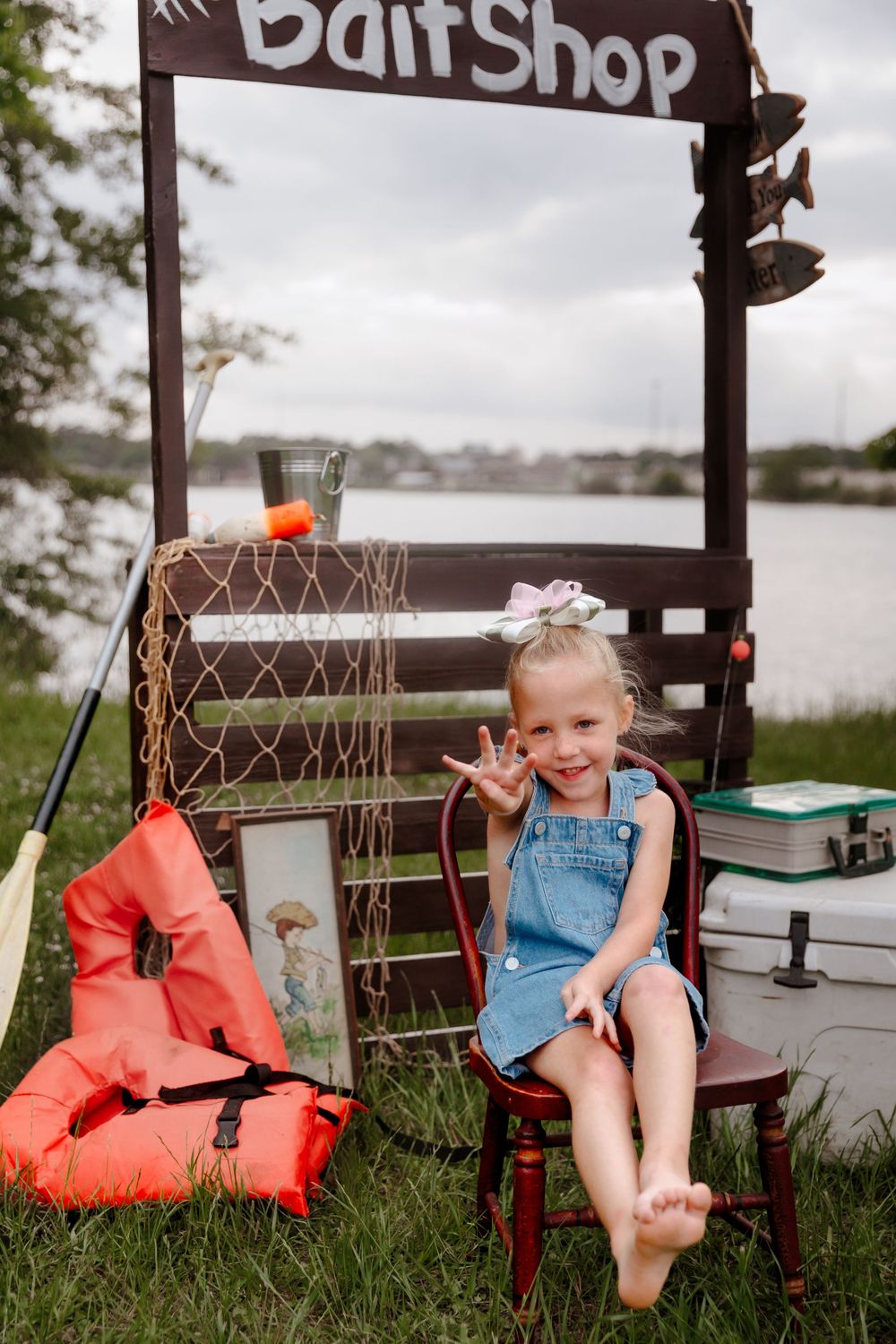 Young girl sitting at a bait shop display by a lake, wearing denim overalls and a bow, smiling outdoors.