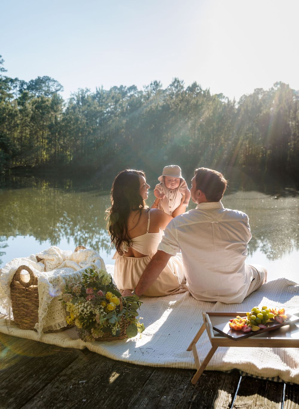 Family sitting on a dock by a serene lake, holding their baby, surrounded by flowers and a picnic spread in golden sunlight.