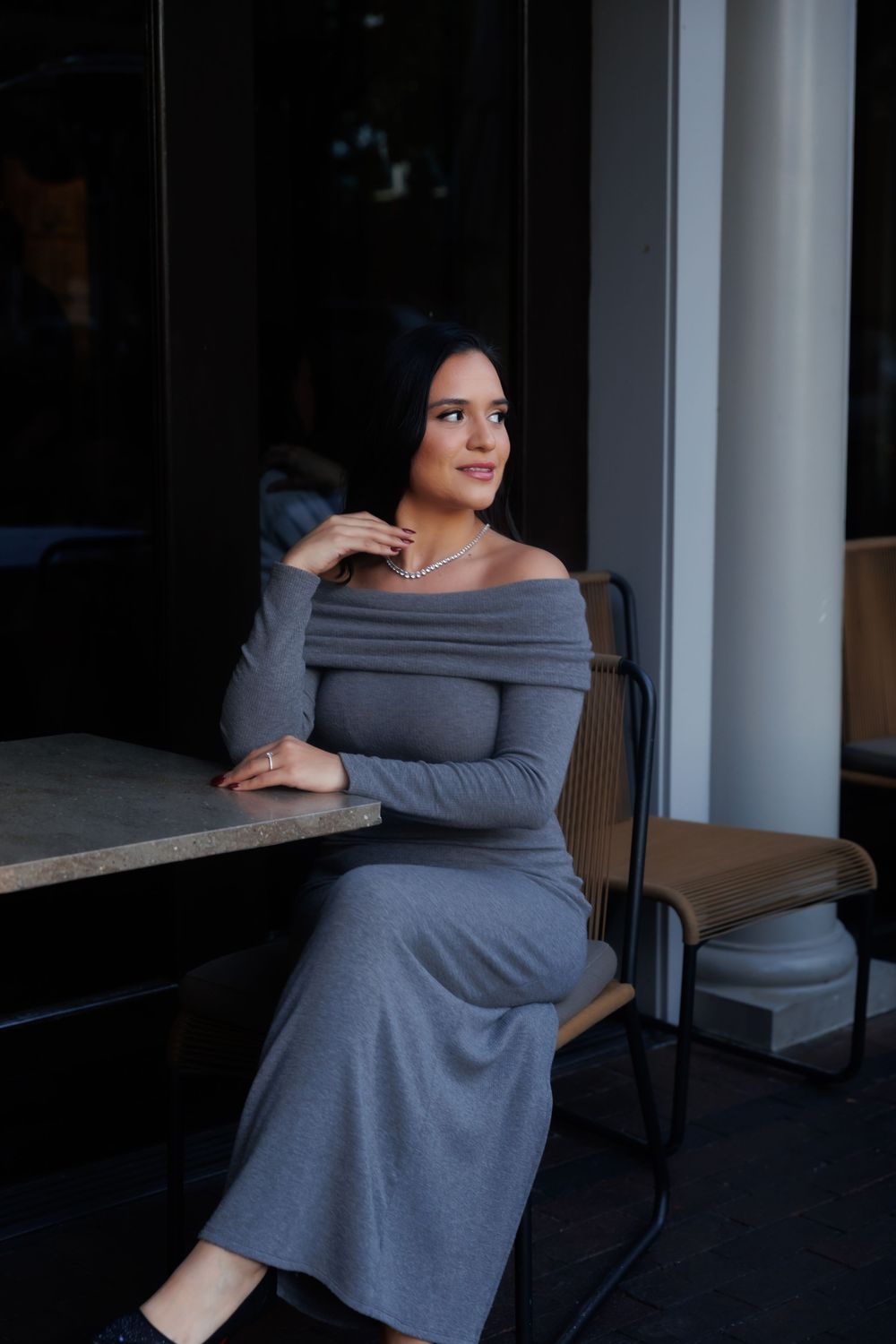 A woman in a gray sweater dress sits elegantly on a chair in a moody, dimly lit setting.