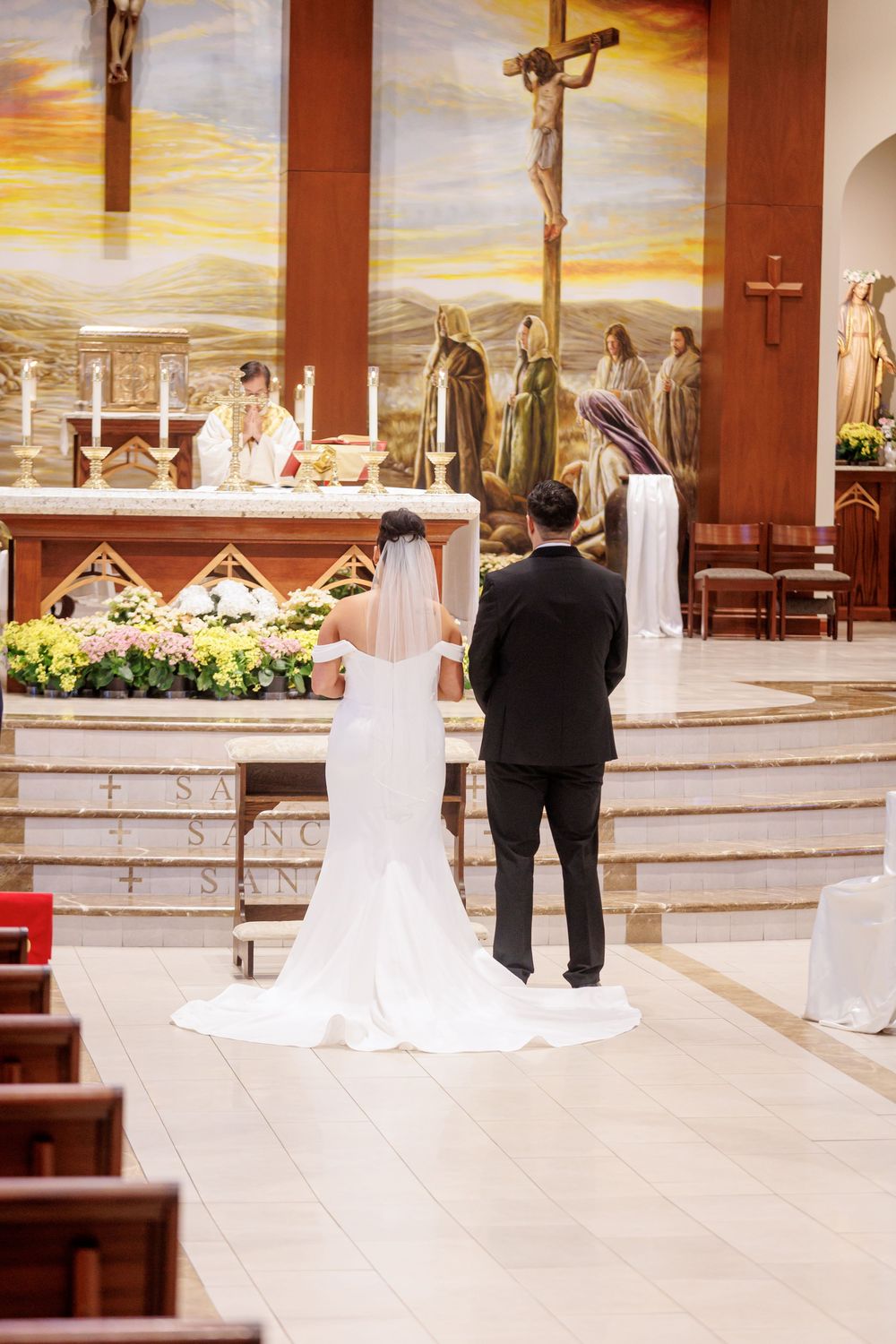 Bride and groom standing at church altar during wedding ceremony with crucifix and floral arrangements in background.