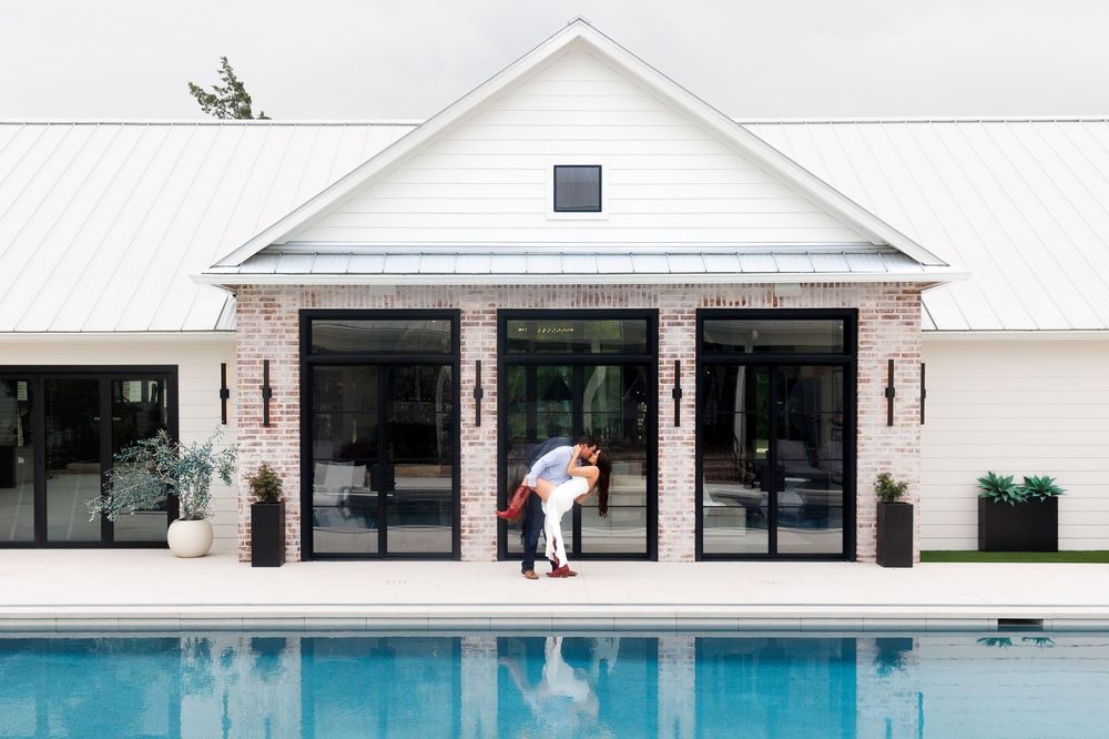 Couple embracing by a modern farmhouse pool with large glass doors, white roof, and brick accents on a sunny day.