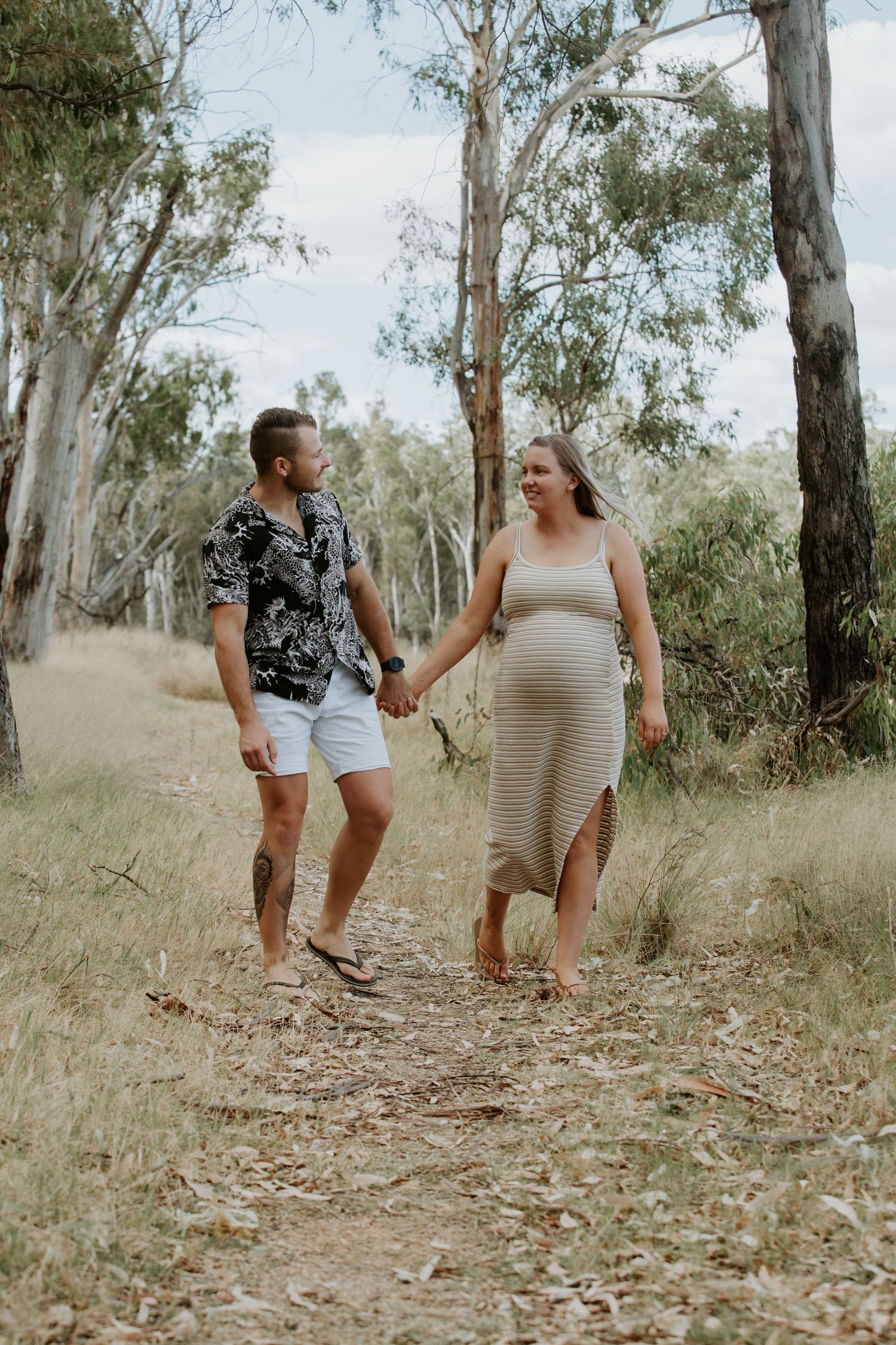 Two people walking and holding hands on a dirt path surrounded by eucalyptus trees in a natural outdoor setting.