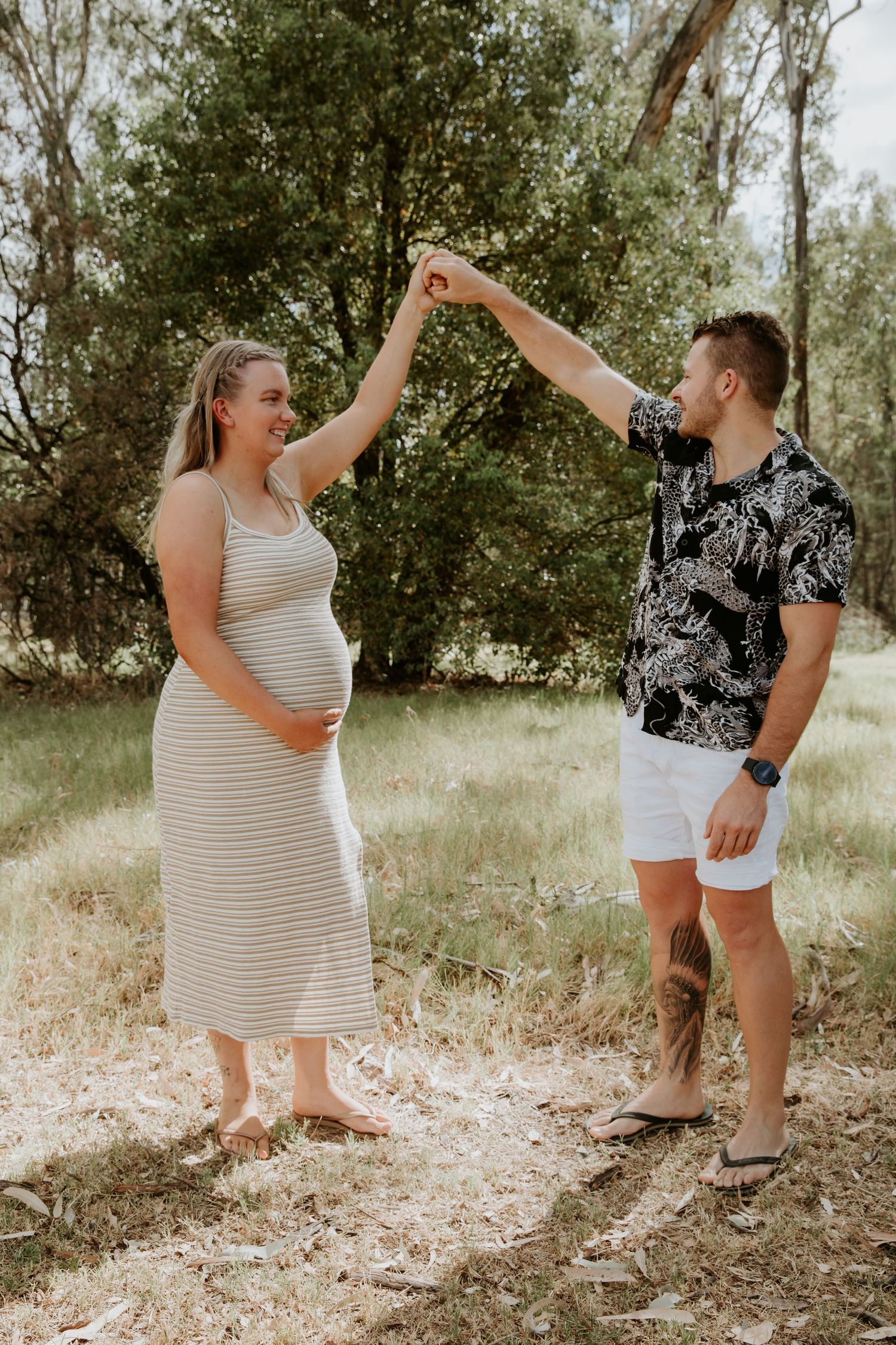 A couple in casual summer outfits playfully dancing and twirling together in a sunny outdoor setting with trees.