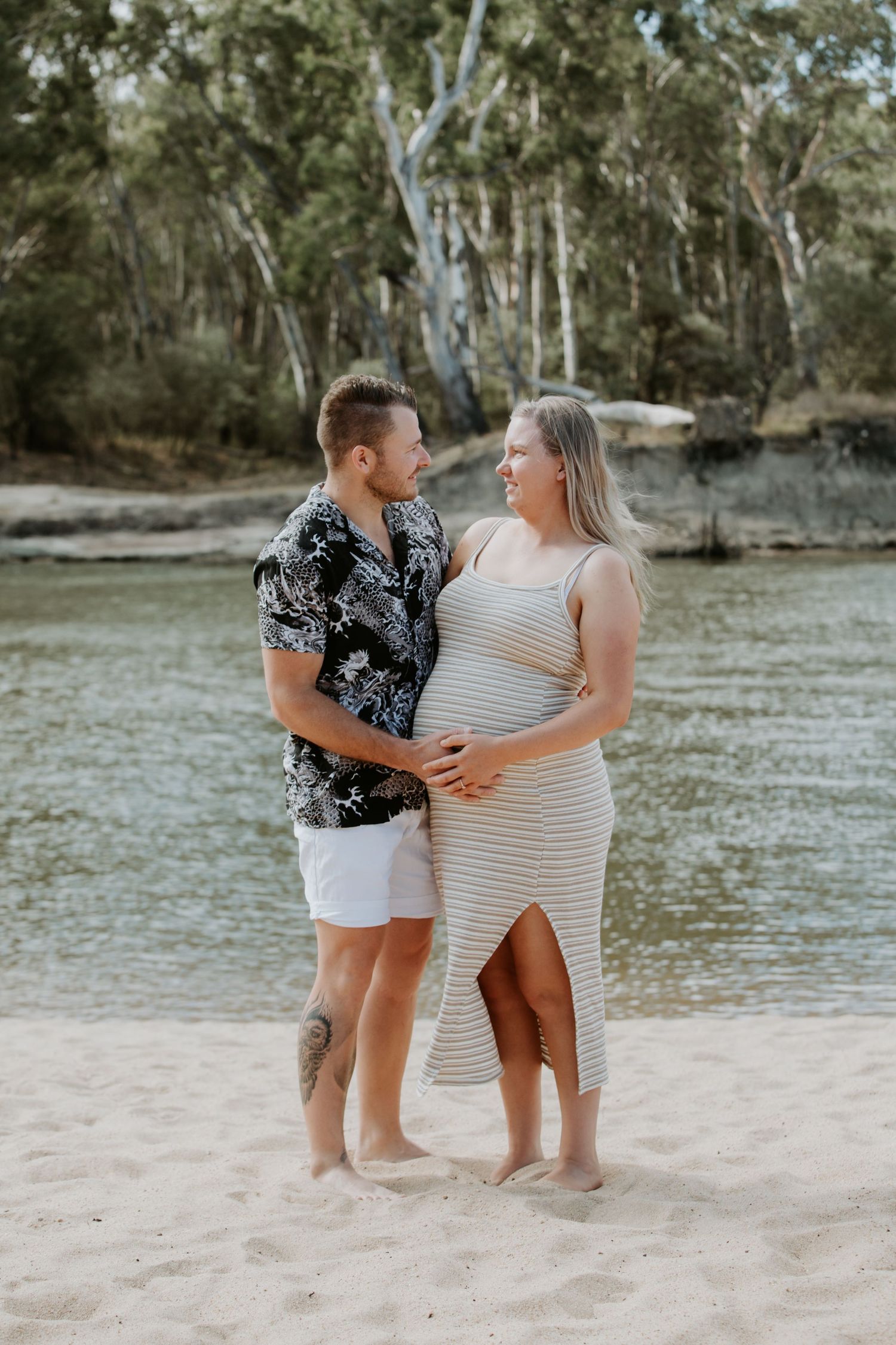 A couple shares a tender moment on a sandy beach with eucalyptus trees in the background.