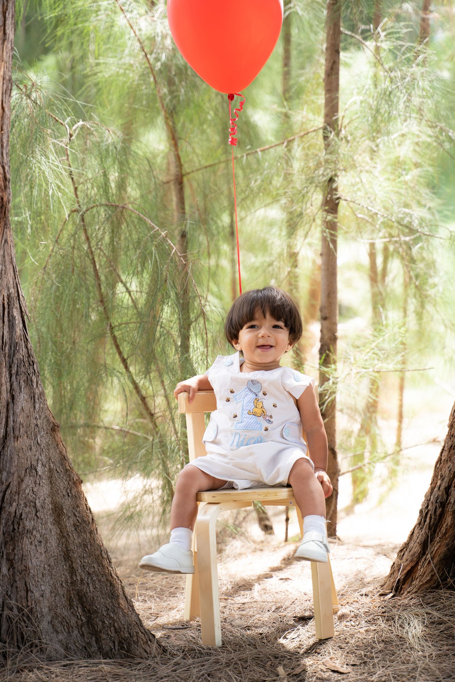 Smiling toddler in white outfit sitting on wooden chair in pine forest with red balloon tied above.