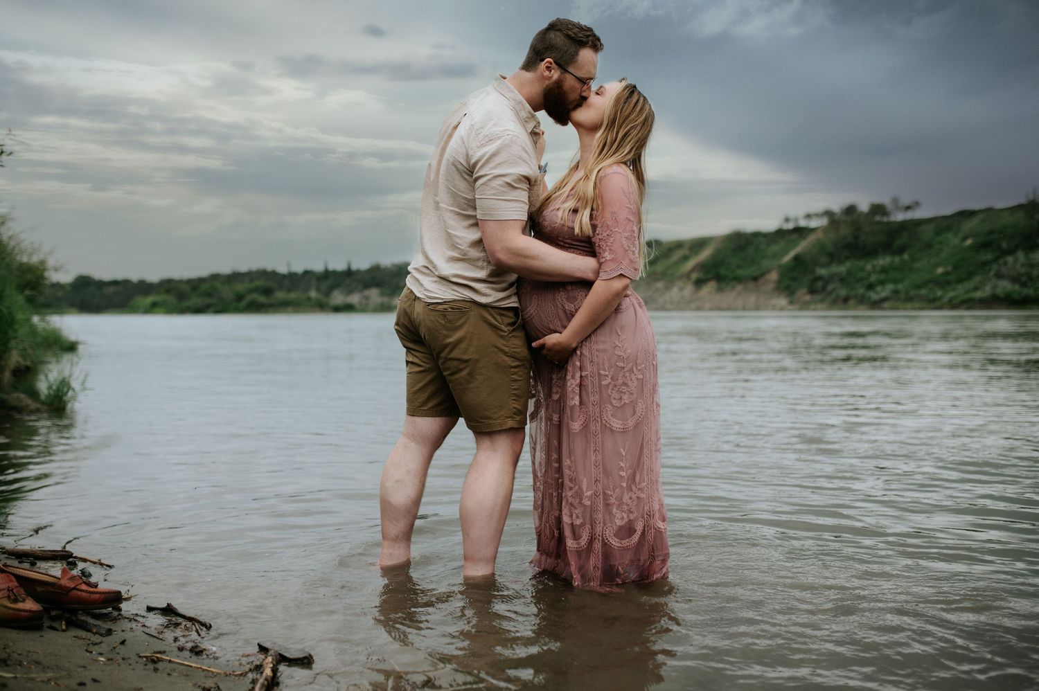 A pregnant woman in a pink lace dress stands on rocks beside a river under dramatic stormy skies.