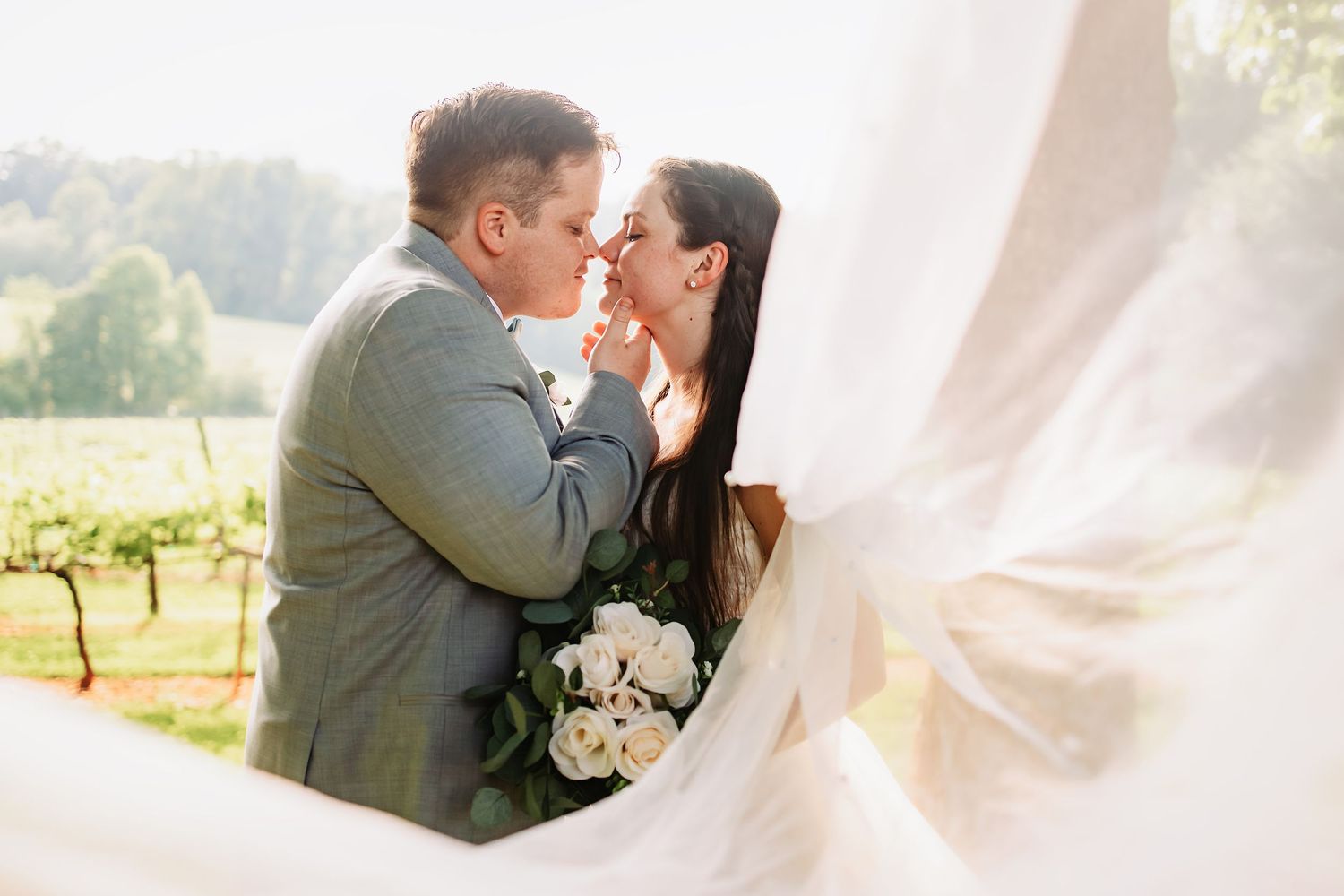 A romantic couple nearly kissing at a vineyard wedding, bride holding white bouquet, veil flowing in the breeze.