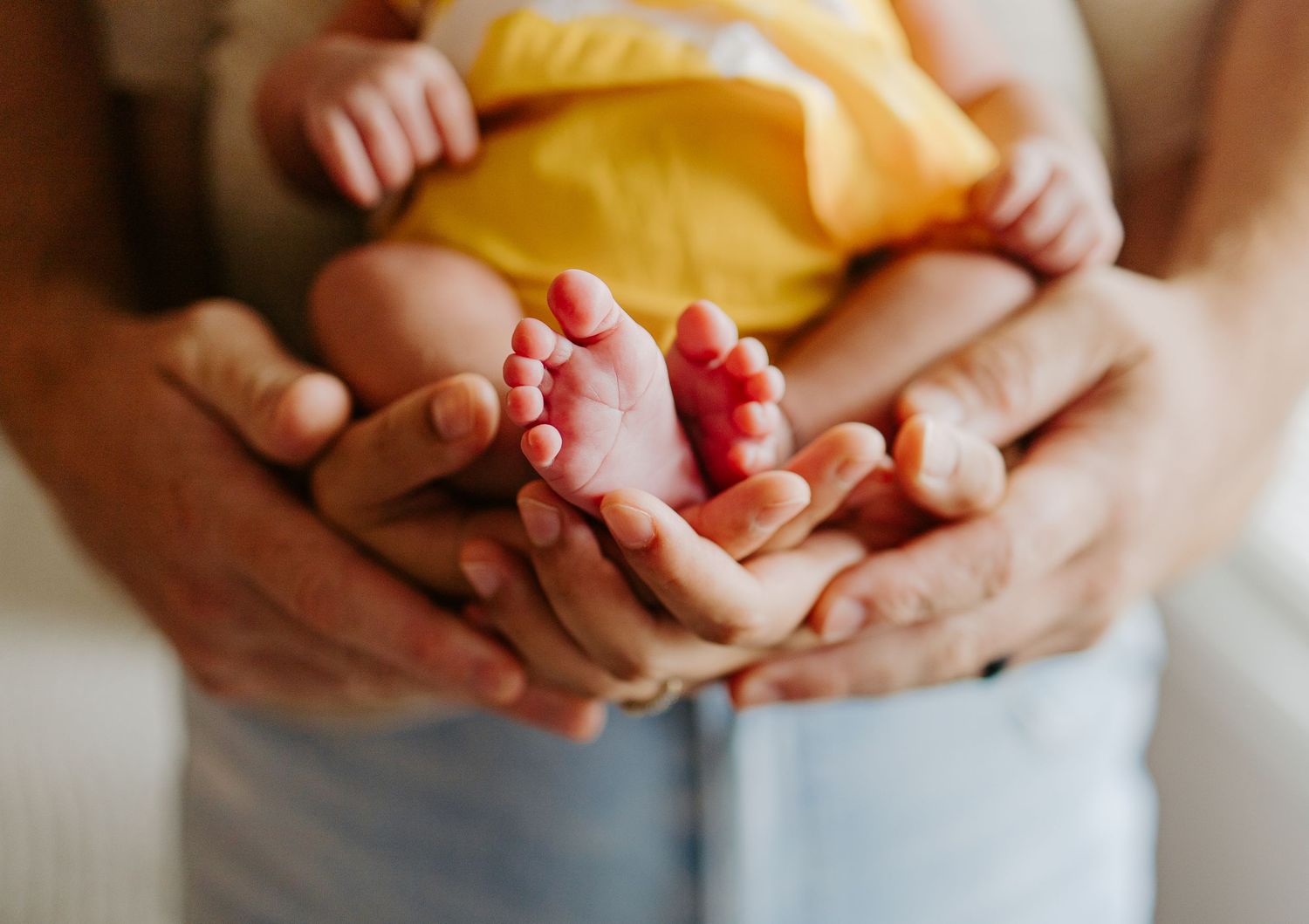 Parents cradle a newborn baby's tiny feet in their hands, baby wearing a yellow outfit in a tender family moment.