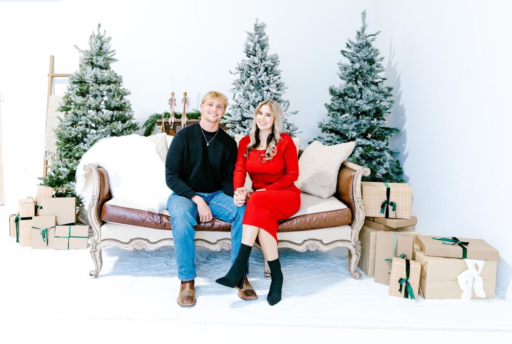 Holiday portrait of couple sitting on vintage sofa with wrapped presents and evergreen trees in studio setting.
