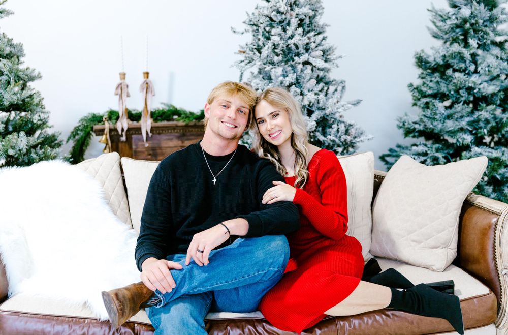 A couple sits together on an elegant white couch with snowy evergreen trees and candlesticks in the background.