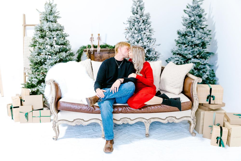 A couple relaxes on a vintage white sofa against a winter backdrop with frosted pine trees and decorative candles.