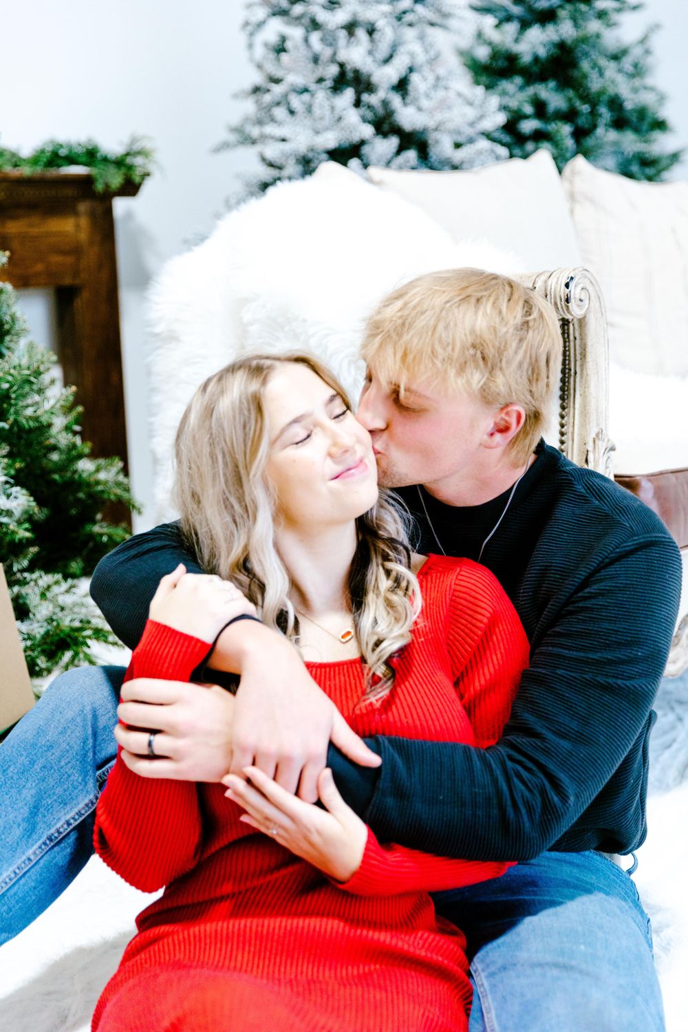 A couple shares a romantic winter moment on a vintage chair with snowy evergreens in the background.