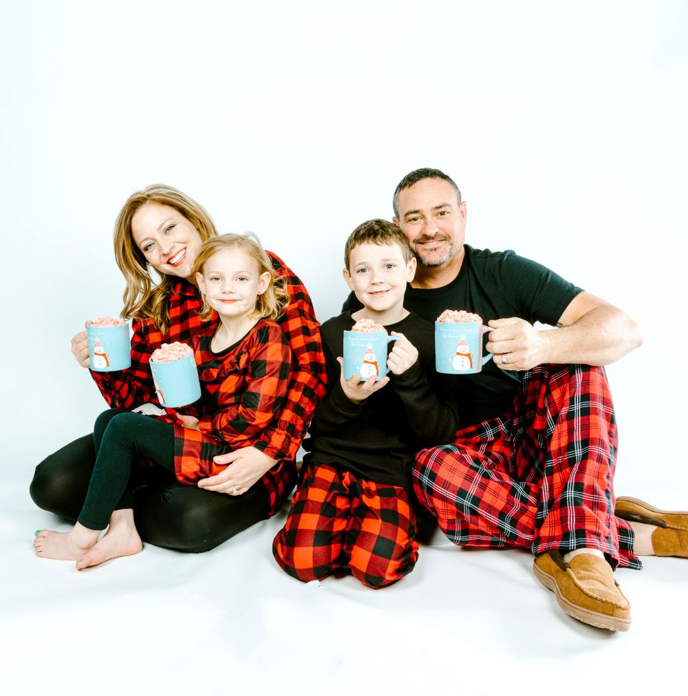 Family wearing matching buffalo plaid pajamas sits together holding blue coffee mugs on white background.