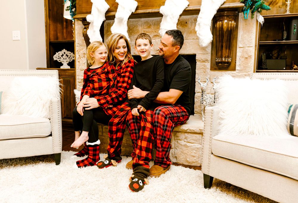 Family wearing matching red and black buffalo plaid pajamas sits together on white couch during holiday season.