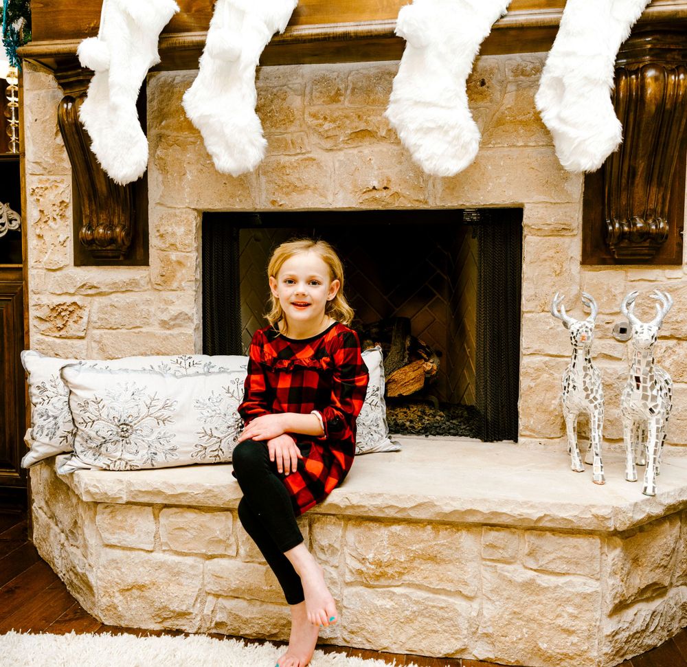 Christmas stockings hang above a stone fireplace with a child sitting on the hearth in buffalo plaid pajamas.