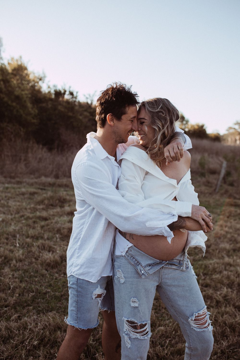 A romantic moment between two people in casual white and denim outfits embracing in a sunlit meadow at dusk.