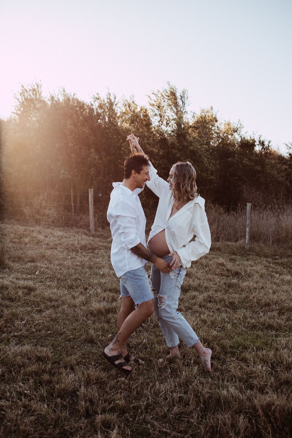 A couple dancing together in a field at sunset wearing casual white outfits and creating a romantic atmosphere.