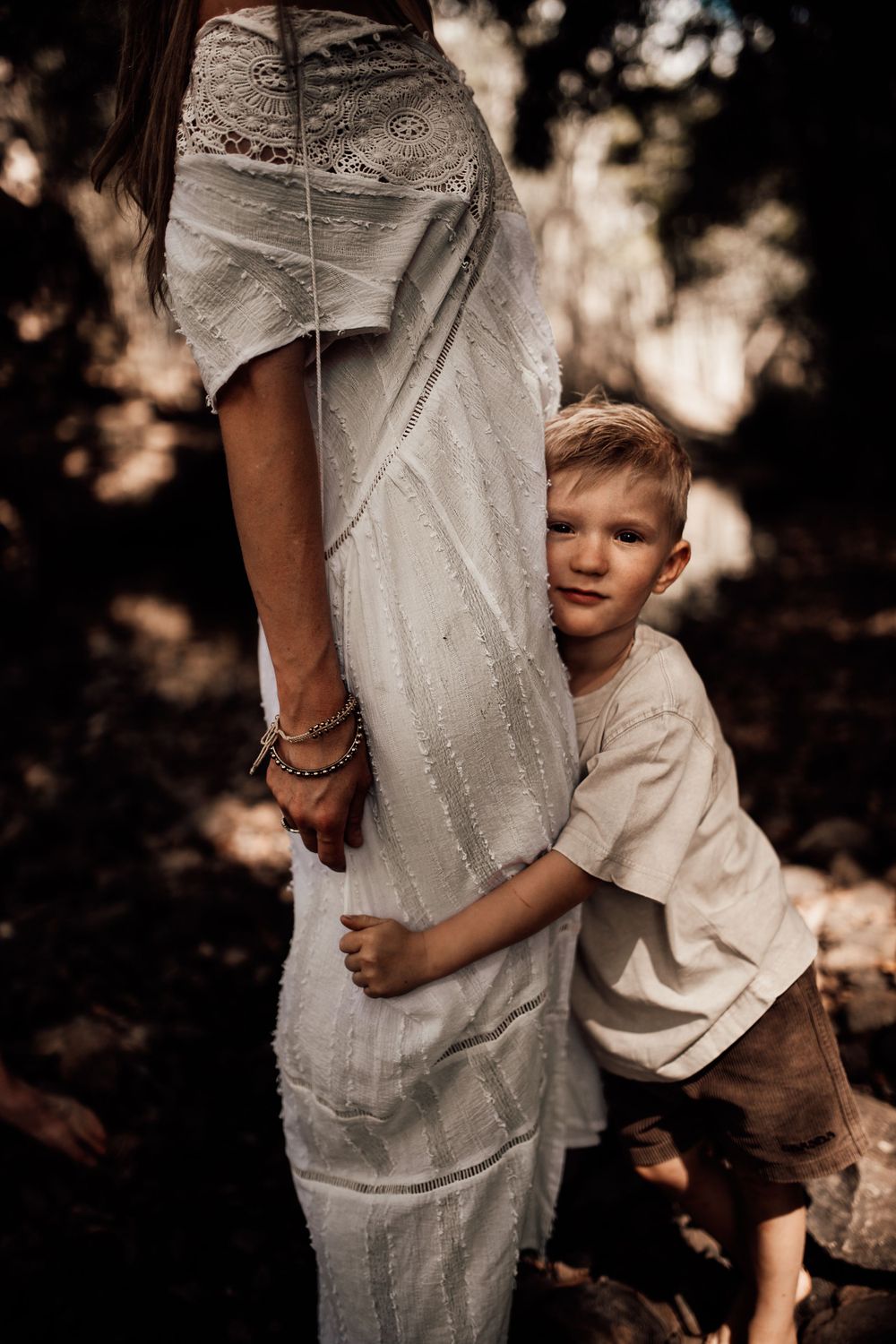 A young child in a white shirt hugs his parent wearing a long white lace dress in a dreamy outdoor setting.