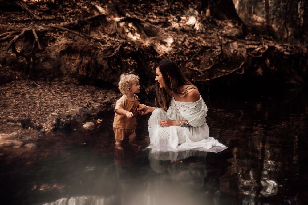 Two figures in white dresses explore a mystical woodland pond with ethereal brown tones in the surrounding nature.