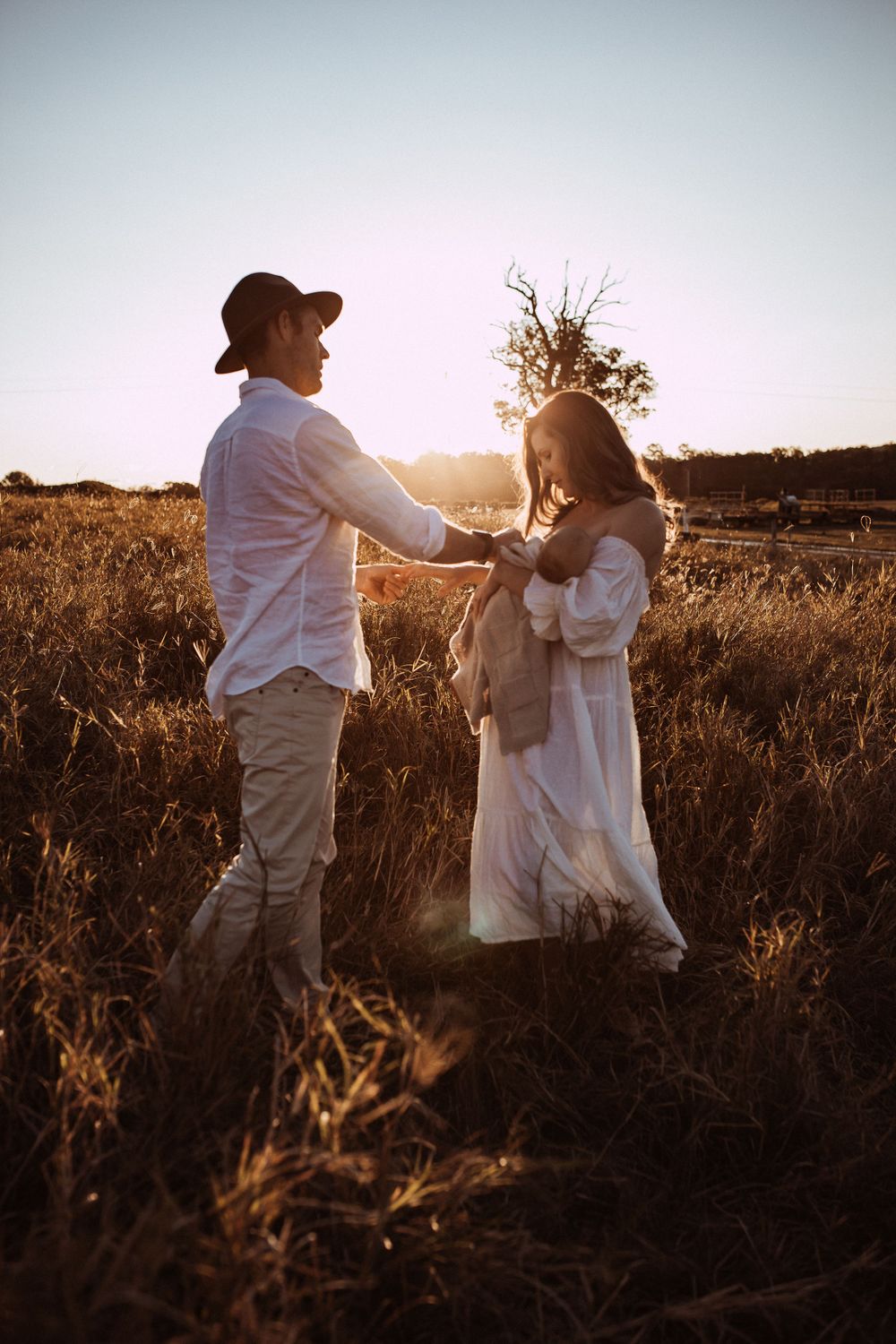 A couple in white clothing dances together in a golden field during sunset while holding hands.