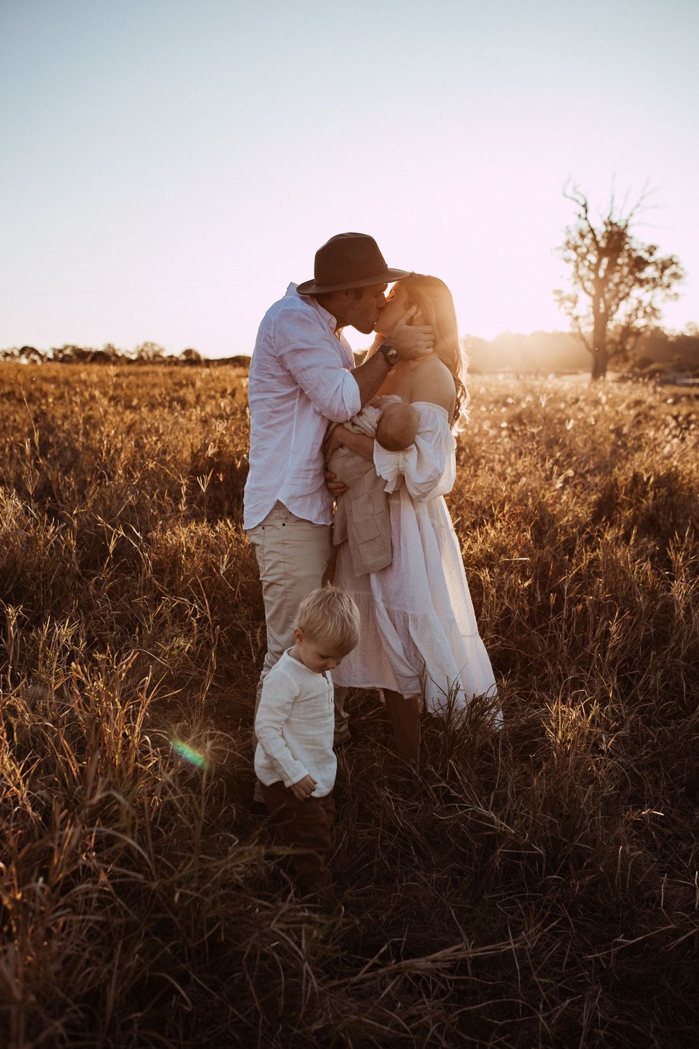 A couple embraces in a golden field at sunset while wearing light colored clothing and a brown hat.