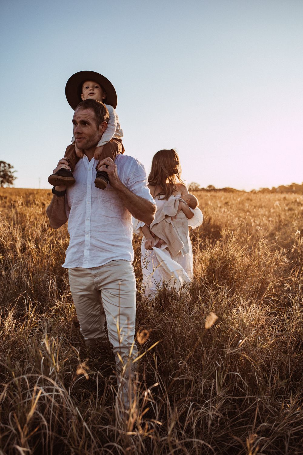 A family walks together through a golden wheat field at sunset, with a child wearing a hat sitting on shoulders.