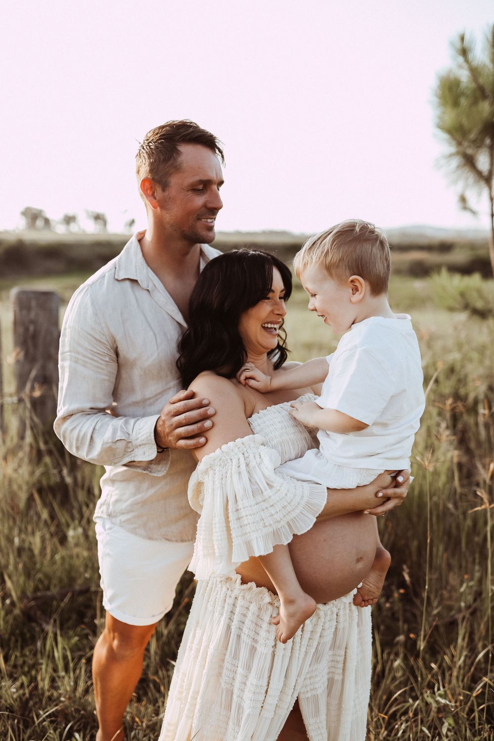 Sweet family moment captured in sunlit field with rustic fence and trees.