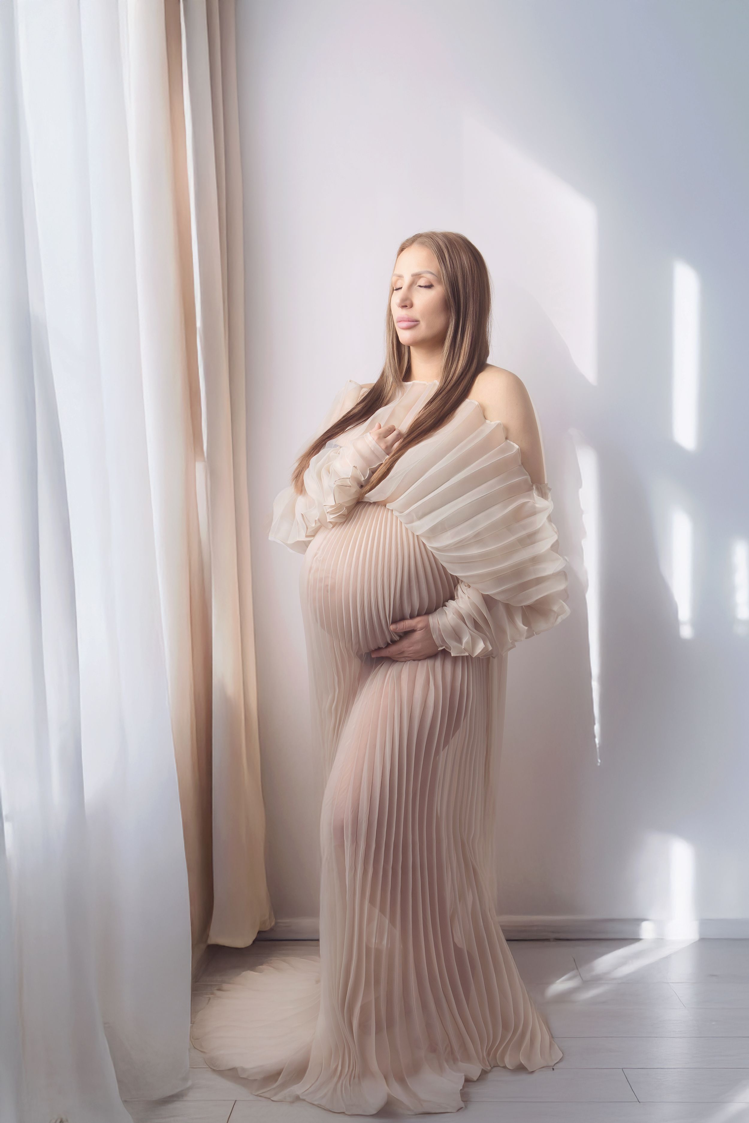 Pregnant woman in beige pleated gown holding baby bump, standing by white curtains in soft natural light.