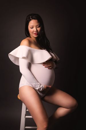 Maternity portrait with subject in white off-shoulder bodysuit seated on stool.