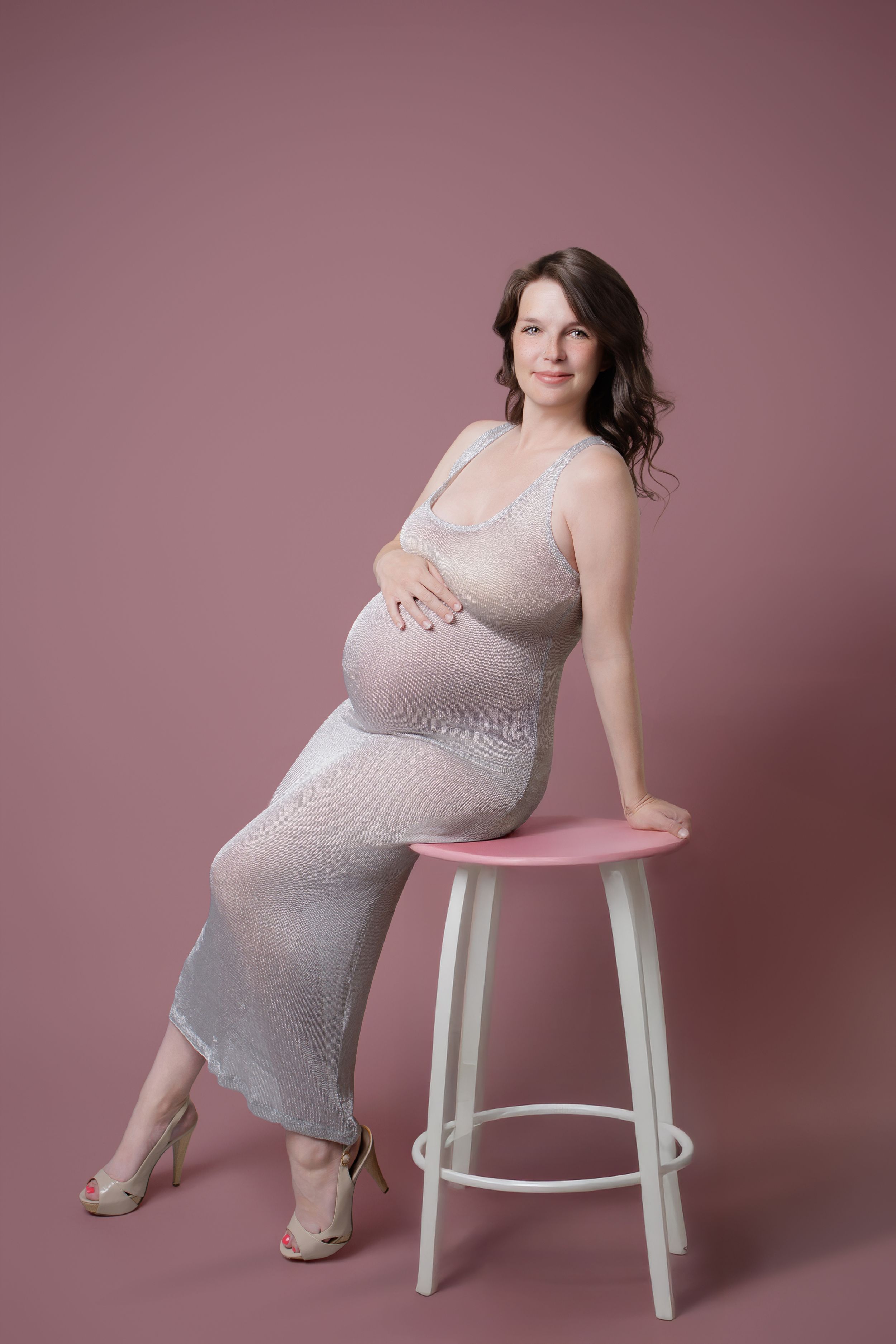 Maternity portrait series in gray dress showing different seated poses against pink backdrop with white stool.